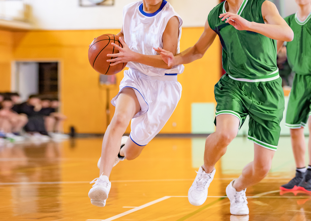 A basketball player in a white uniform drives past defenders dressed in green uniforms during a game at a gym.