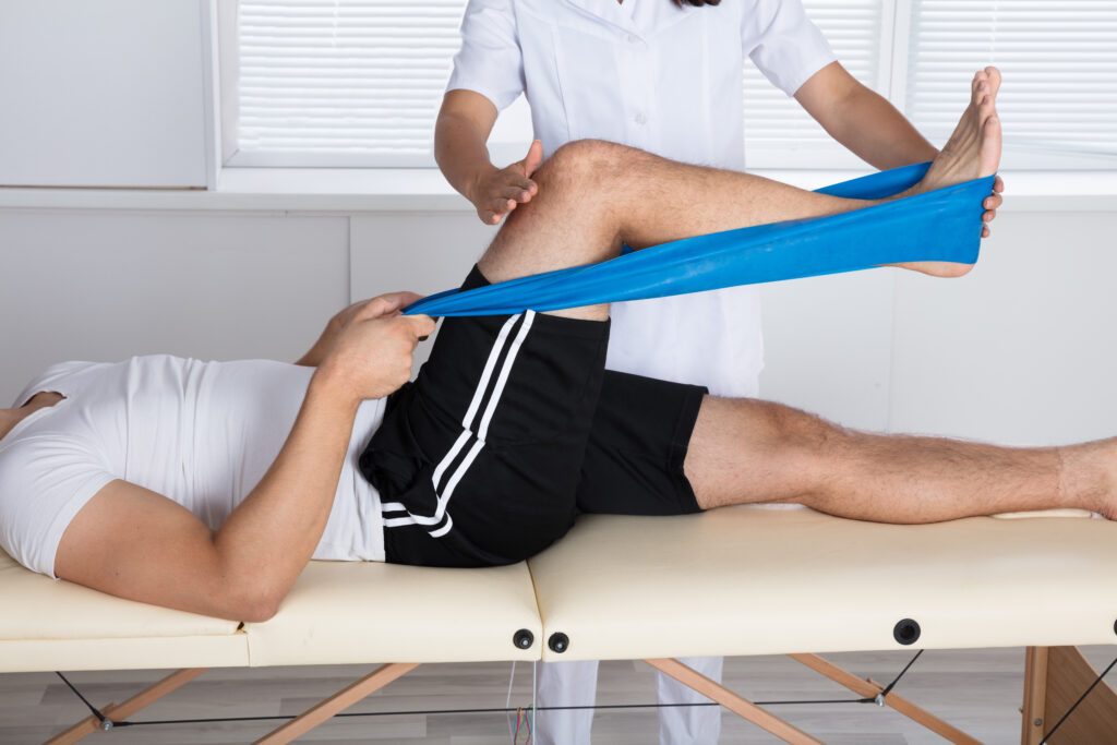 A patient stretches out during a physical therapy session. They are using an elastic band to stretch their leg with the assistance of a physical therapist.